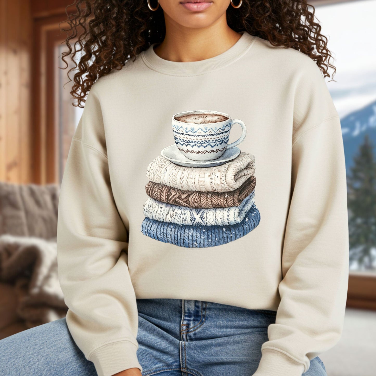 Woman in a cream sweatshirt featuring a watercolor stack of knit sweaters and a patterned coffee mug.