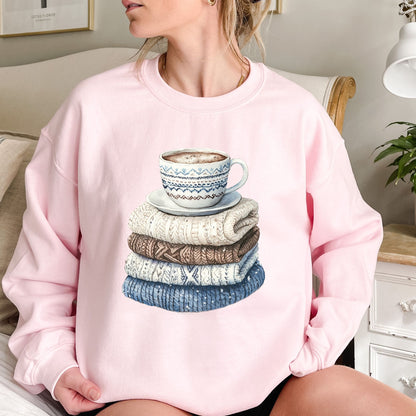 Pink sweatshirt with a watercolor-style print of stacked knit sweaters and a patterned coffee mug.