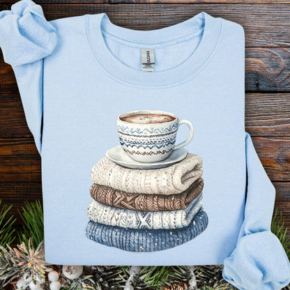 Light blue Gildan sweatshirt with a watercolor print of a patterned mug on a stack of knit sweaters.
