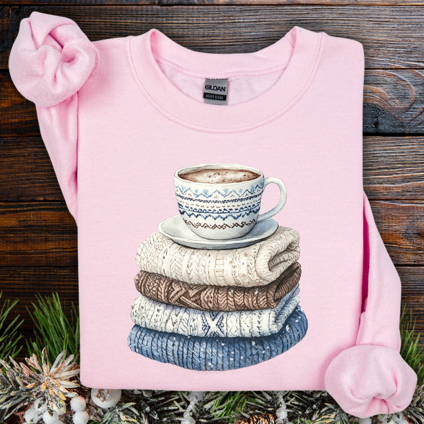 Light pink Gildan sweatshirt with a watercolor graphic of a patterned mug on a stack of folded knit sweaters.