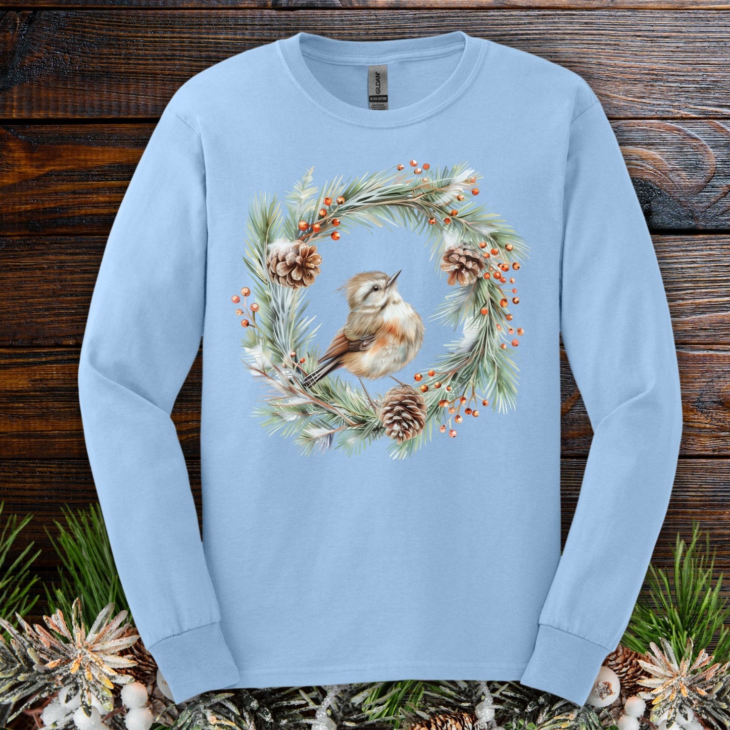 Light blue long sleeve tee with a watercolor winter wren in an evergreen wreath with pinecones and berries.