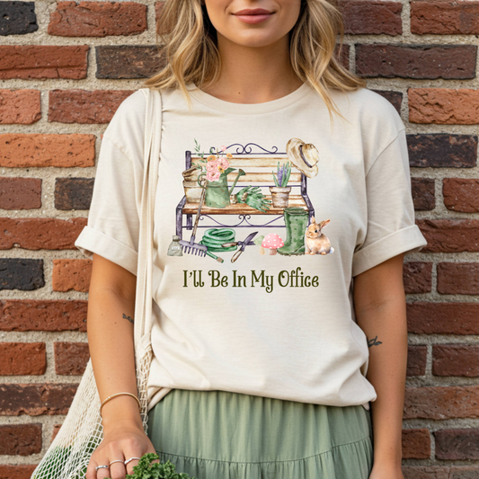 woman wearing a t-shirt with a garden-themed design and text, standing against a brick wall.